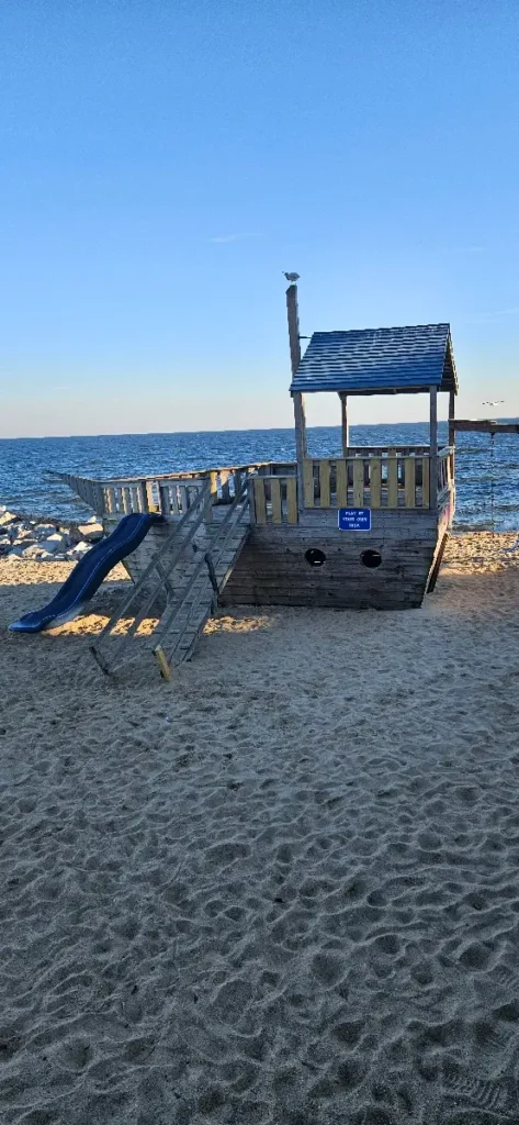 Wooden children's play structure designed to resemble a ship with a slide and a raised deck on a sandy beach. The ocean is visible in the background under a clear blue sky.