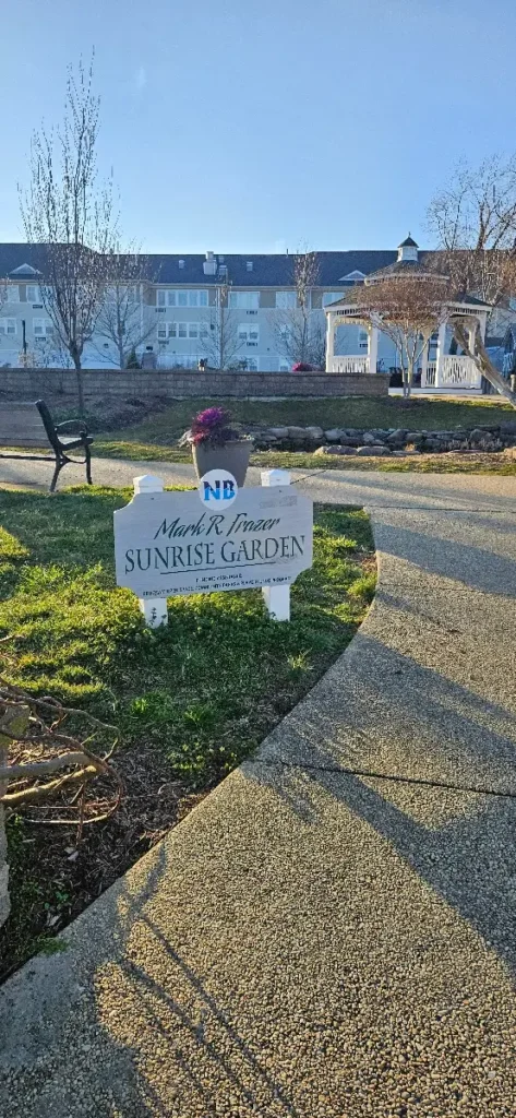 A white sign with 'Mark R. Frazer Sunrise Garden' written on it, in a landscaped area with a concrete path leading towards a gazebo and a multi-story building in the background. There are leafless young trees and a clear blue sky above.
