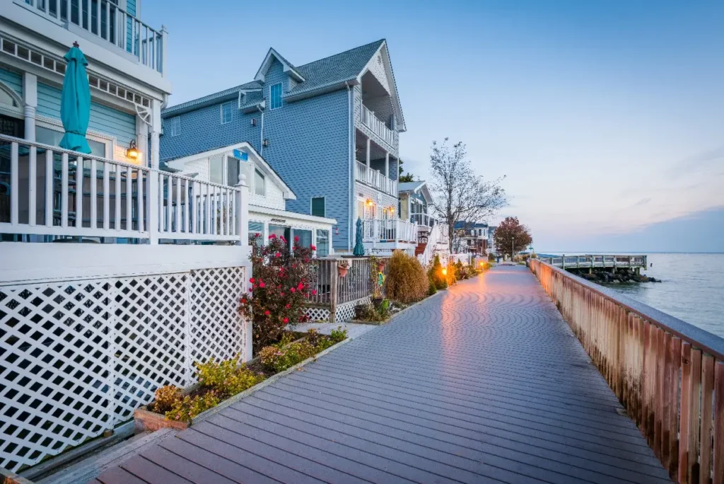 A picturesque boardwalk illuminated by warm street lamps at twilight. The walkway is flanked by charming blue houses with white railings and decorative lattices. Lush rose bushes and other greenery add color to the scene, and a quiet bay stretches into the distance, with a peaceful sky overhead.